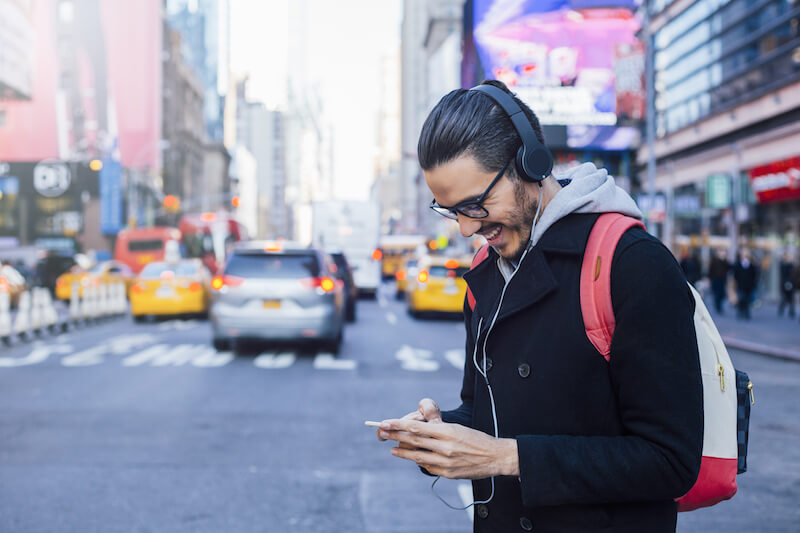 Man wearing headphones on a street smiling at a mobile phone.