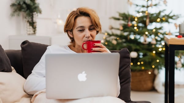 A person holding a mug whilst looking at a laptop, with a Christmas tree in the background.