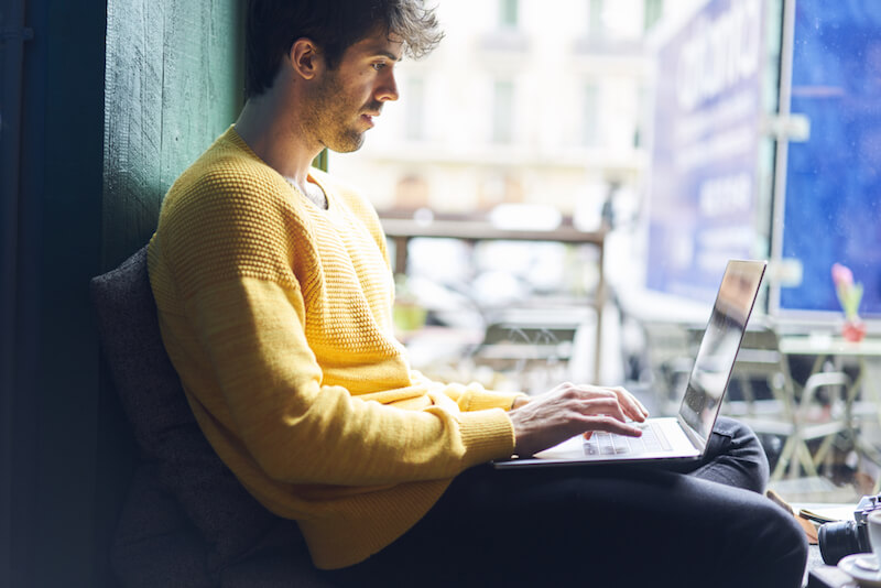 man sitting beside window wearing yellow jumper typing into laptop