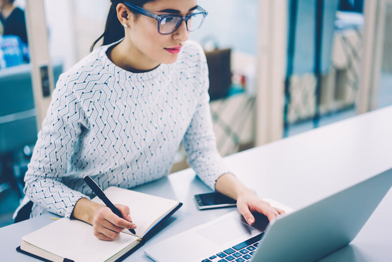 woman wearing glasses working at laptop biting a pencil